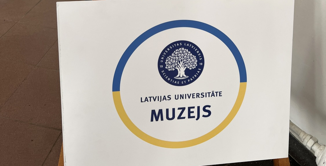 Stends ar Latvijas Universitātes Muzeja nosaukumu un LU emblēmu centrā, ko ieskauj apaļš zils un dzeltens aplis uz balta fona.